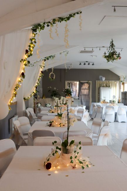 A wedding reception venue decorated with elegant floral arrangements and string lights. The tables are draped with white tablecloths and feature vases with white flowers, complemented by small fairy l