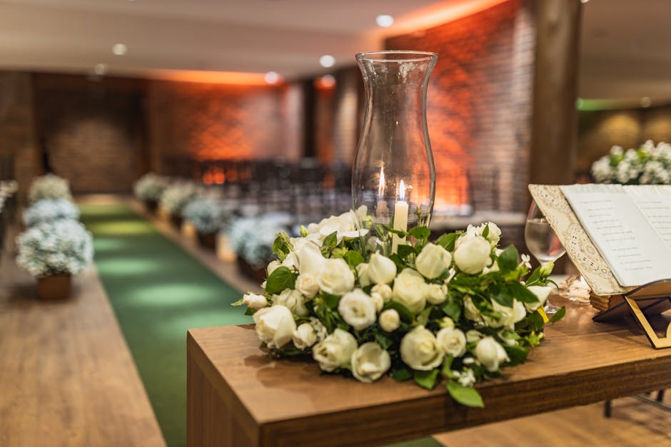 A floral arrangement featuring a bouquet of fresh white roses with lush green foliage, placed on a wooden table next to a tall, clear glass vase and an open music score. The roses are in full bloom wi