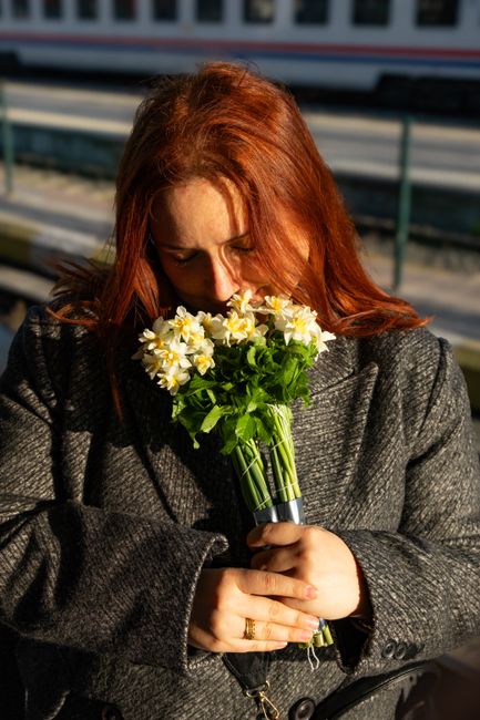 A woman with long red hair holds a bouquet of fresh, white and yellow daffodils with green foliage, wrapped with a simple clear plastic cover. She is dressed in a dark grey, textured coat and appears 