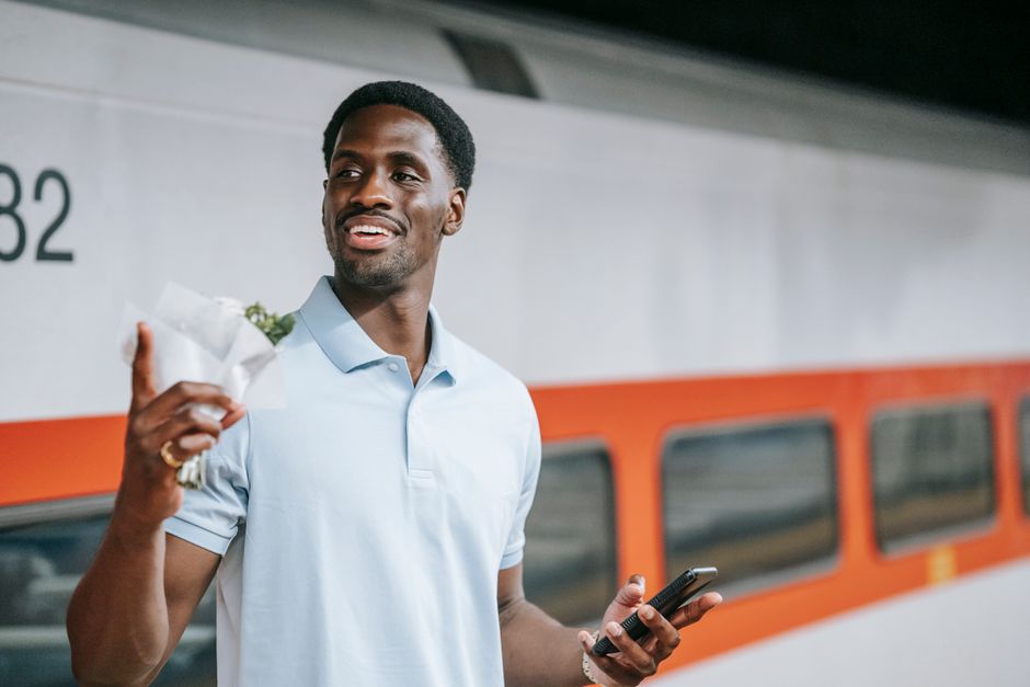 A smiling man holding a bouquet of fresh flowers wrapped in white paper stands beside a train at Crayford railway station. The bouquet includes a variety of flowers with vibrant colors, possibly roses