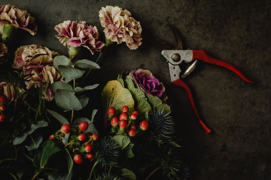 A floral arrangement featuring pink and cream carnations, purple ornamental cabbages, red hypericum berries, and dark green foliage such as eucalyptus leaves, all laid out on a dark, textured surface.