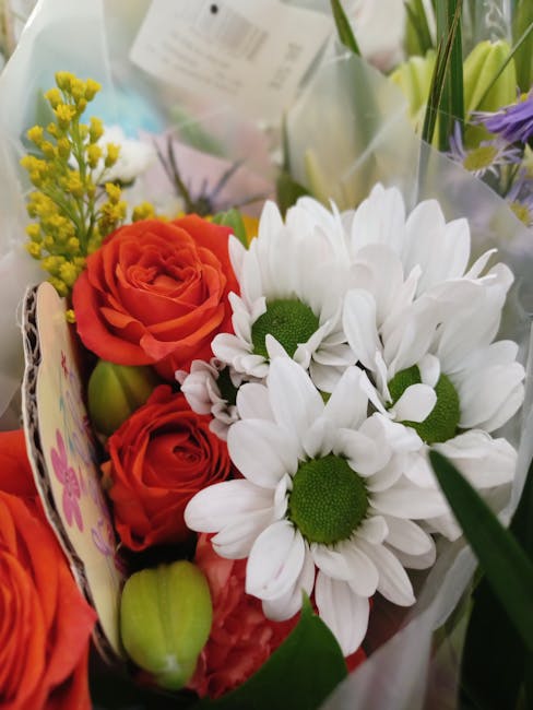 A close-up of a floral arrangement from Crayford Flowers, featuring a vibrant bouquet with orange roses, white daisies with green centers, and sprigs of yellow filler flowers, all wrapped in clear cel